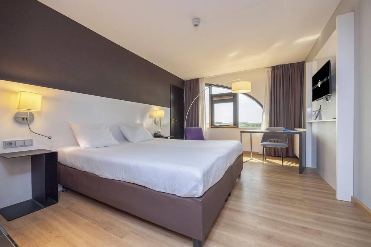 A modern hotel room with a large bed, bedside lamps, a desk with a chair, a TV, and a window with curtains.