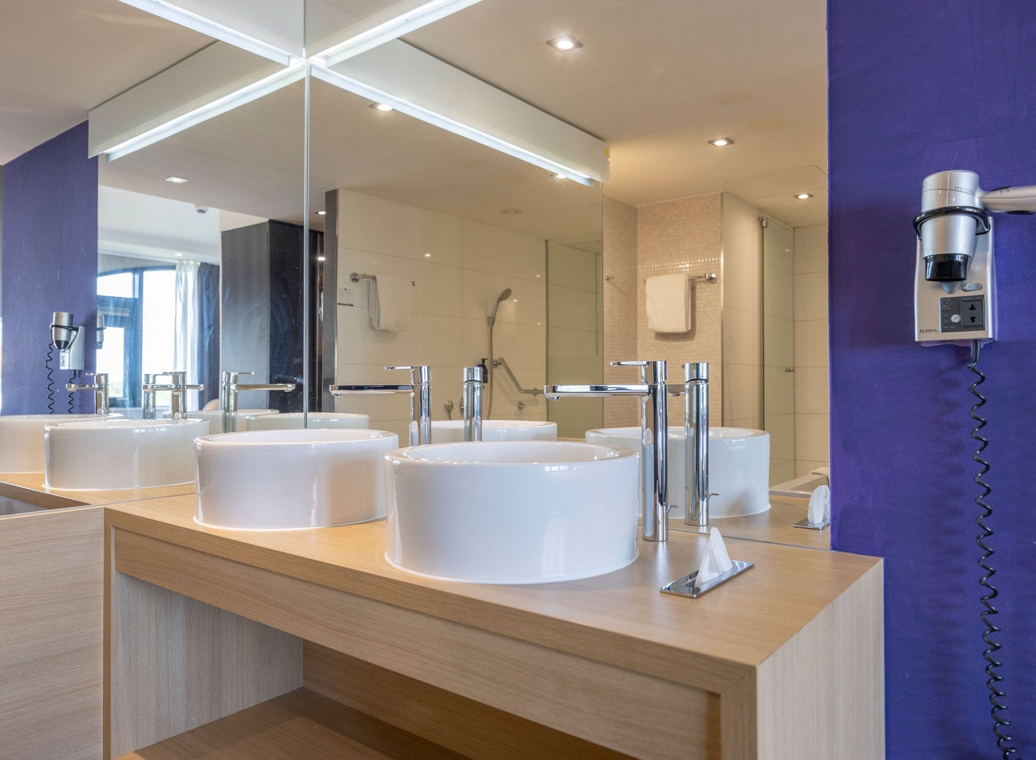 A modern bathroom with three white sinks, a large mirror, and a hairdryer mounted on the wall.