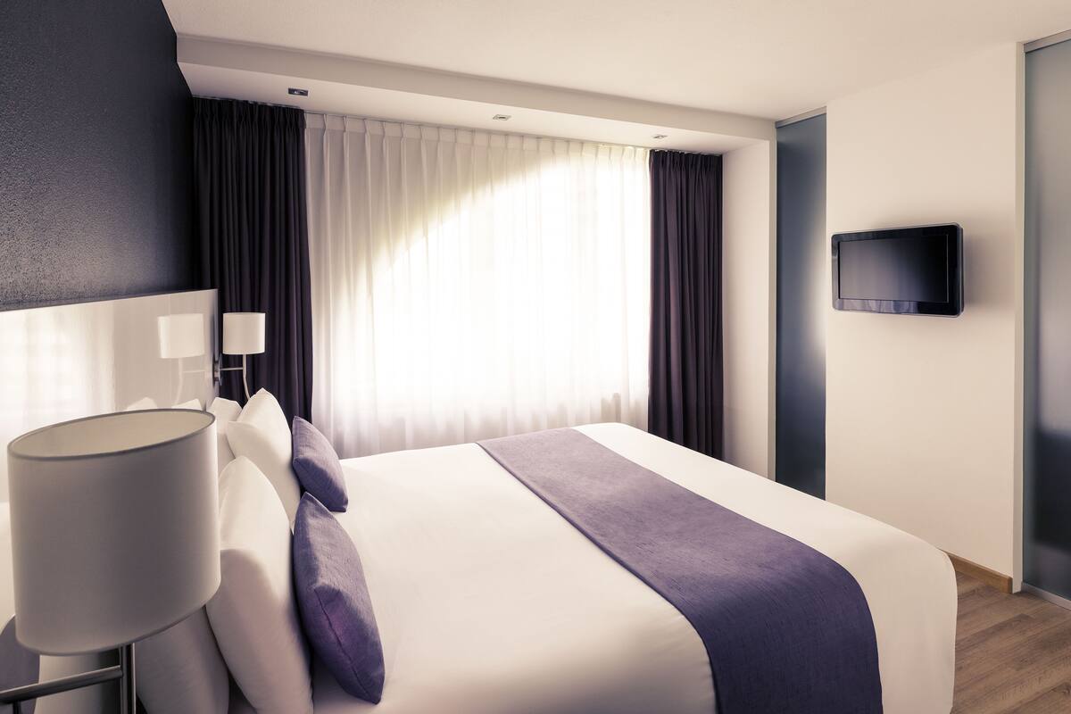 A modern hotel room with a large bed, a bedside lamp, a flat-screen TV mounted on the wall, and a window with curtains.