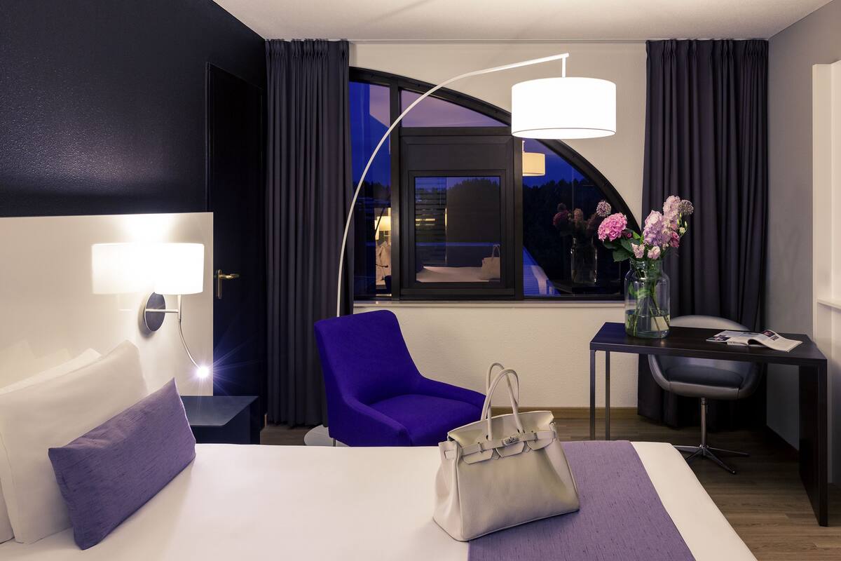 A hotel room with a bed, a desk, a chair, a lamp, and a window with curtains.