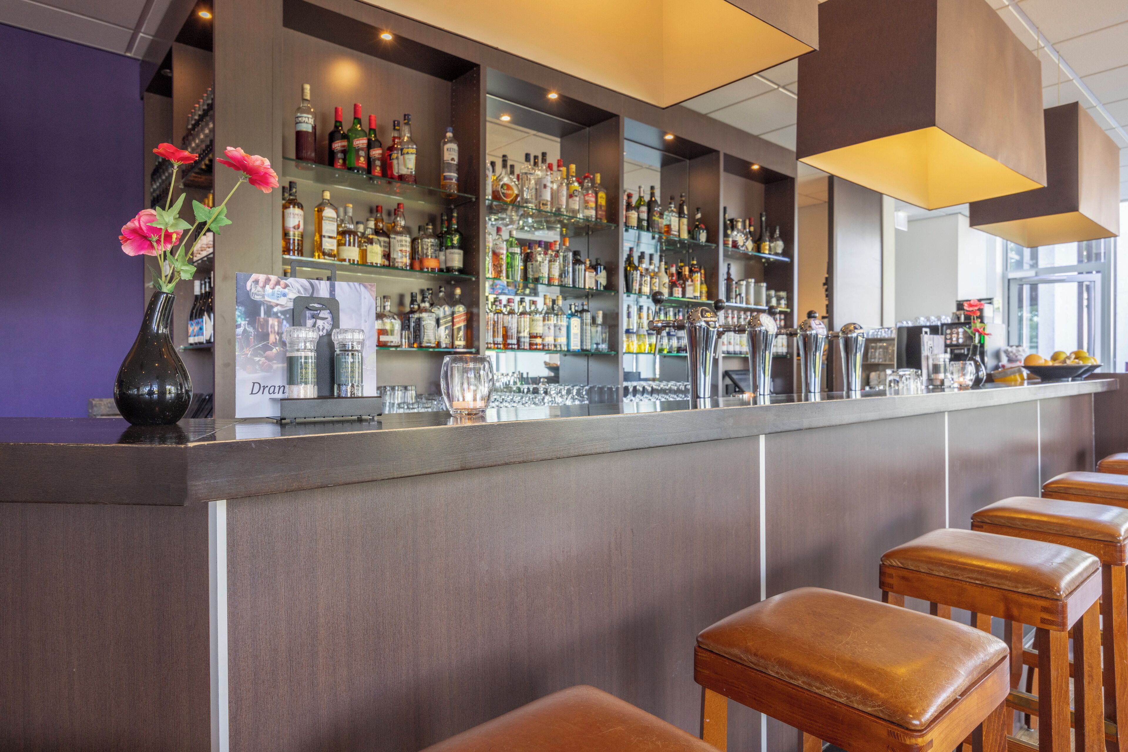 Una zona de bar con una barra larga, varias estanterías llenas de botellas, un jarrón con flores y taburetes de madera.