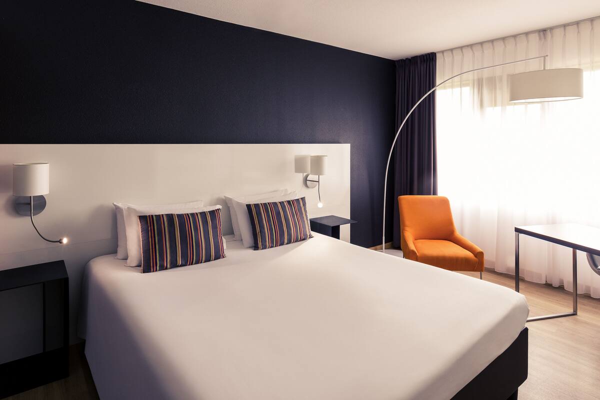 A hotel room with a large bed, two bedside tables, a nightstand, an orange armchair, and a desk with a lamp.