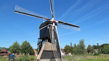 A windmill with a large sail and a bell at the base, surrounded by greenery and houses in the background.