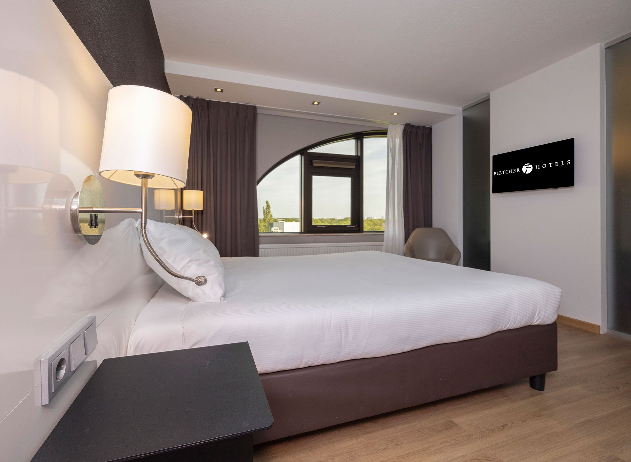 Una habitación de hotel moderna con una cama grande, una mesita de noche, una lámpara, vistas al exterior a través de una ventana y un cartel de FLECHER HOTELS.