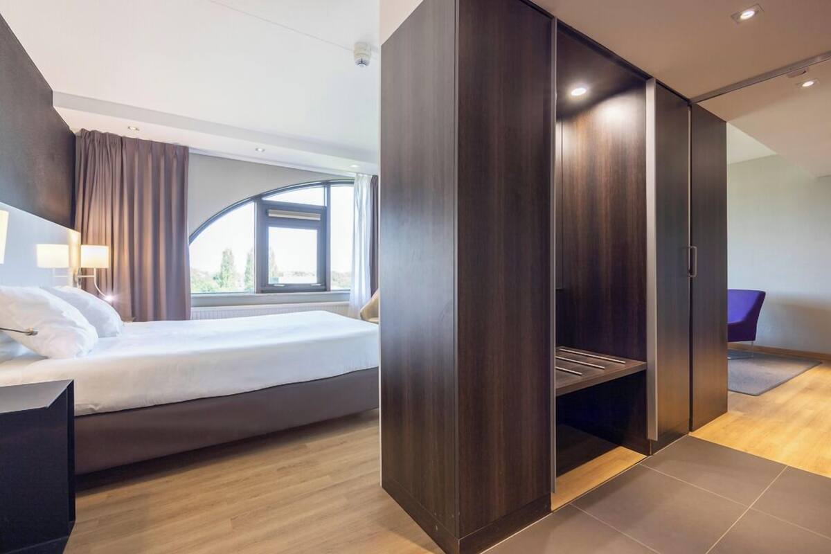 A modern hotel room with a bed, a desk, a wardrobe, and a view of the outdoors.