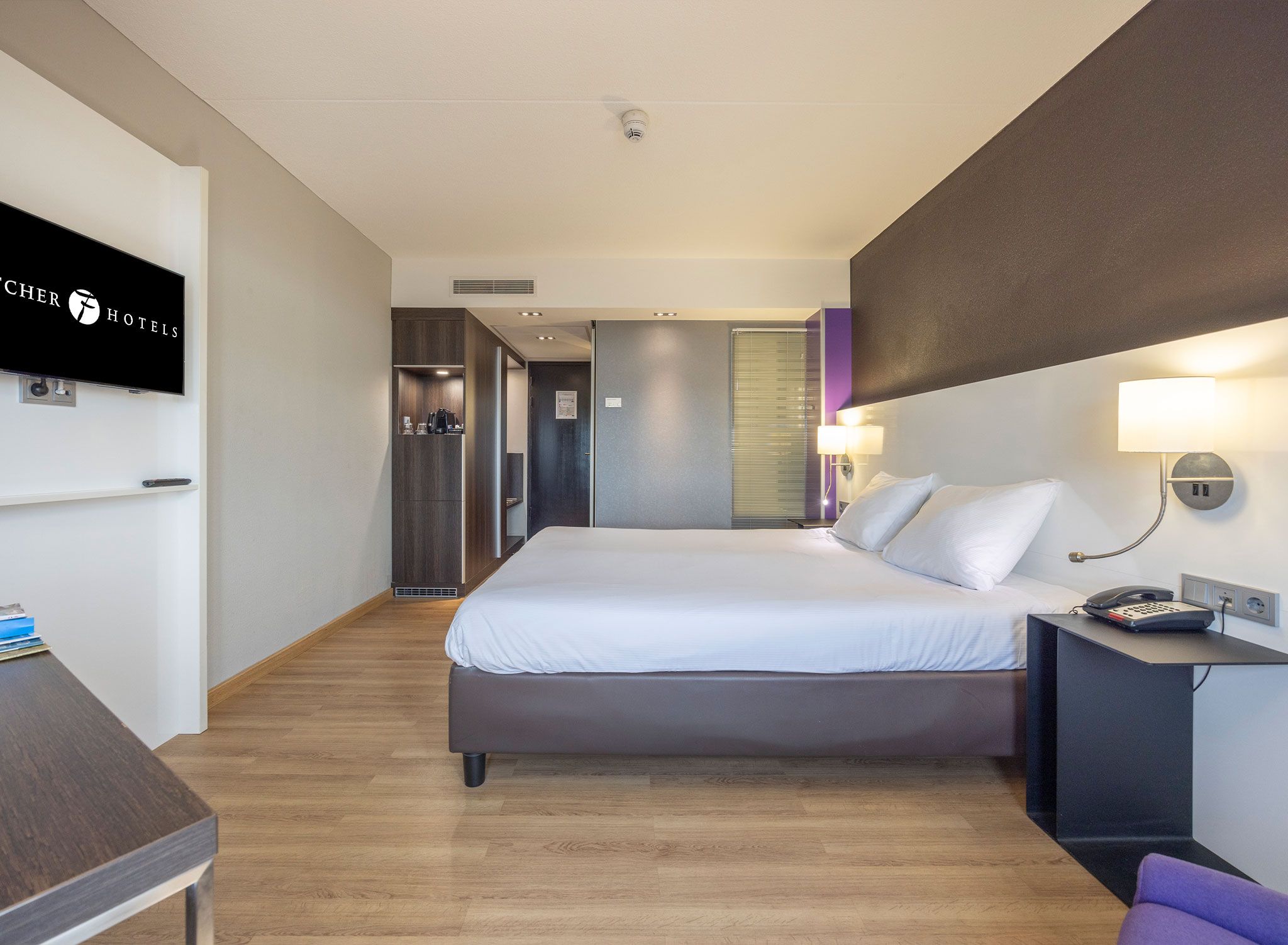 A hotel room with a bed, bedside table, lamp, and a TV mounted on the wall.
