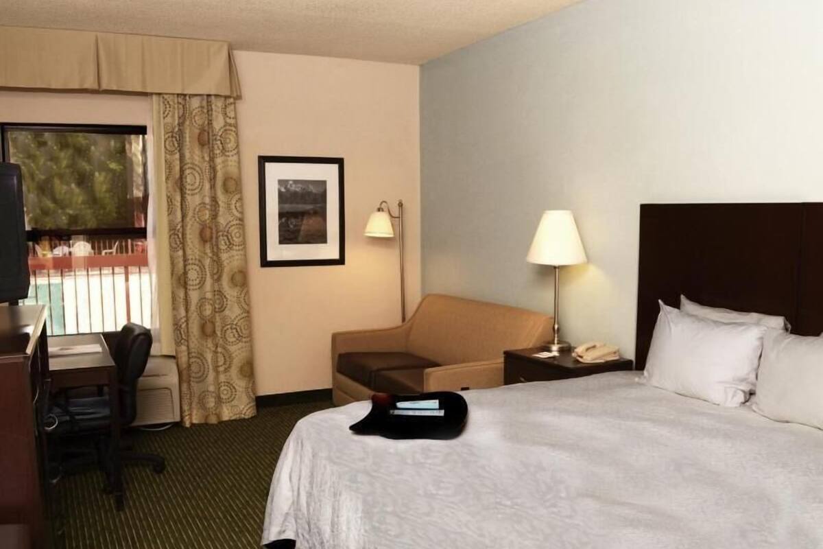 Ein Hotelzimmer mit einem Bett, einem Schreibtisch mit Stuhl, einer Lampe, einem gerahmten Bild an der Wand und einem Fenster mit Vorhängen.