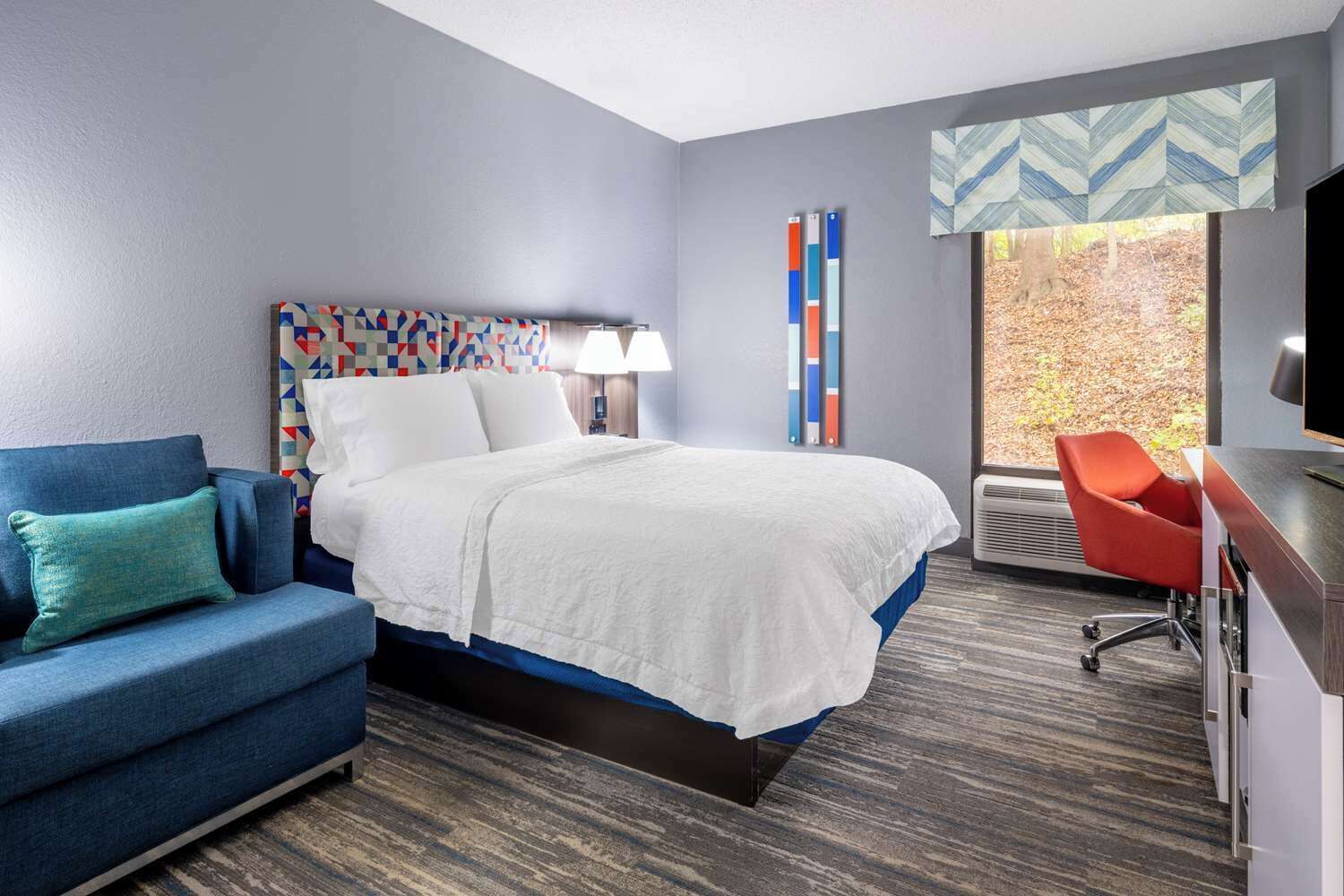 A hotel room with a bed, a blue sofa, a desk, and a window with a view of trees.