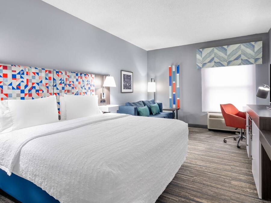 A modern hotel room with a large bed, a desk, a chair, and a TV.