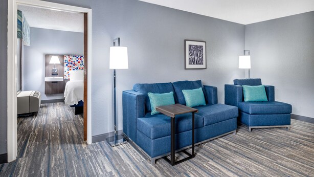 A hotel room with a blue sofa set, two side tables, a patterned carpet, and a framed picture on the wall. - Hampton Inn Sanford (Sanford)