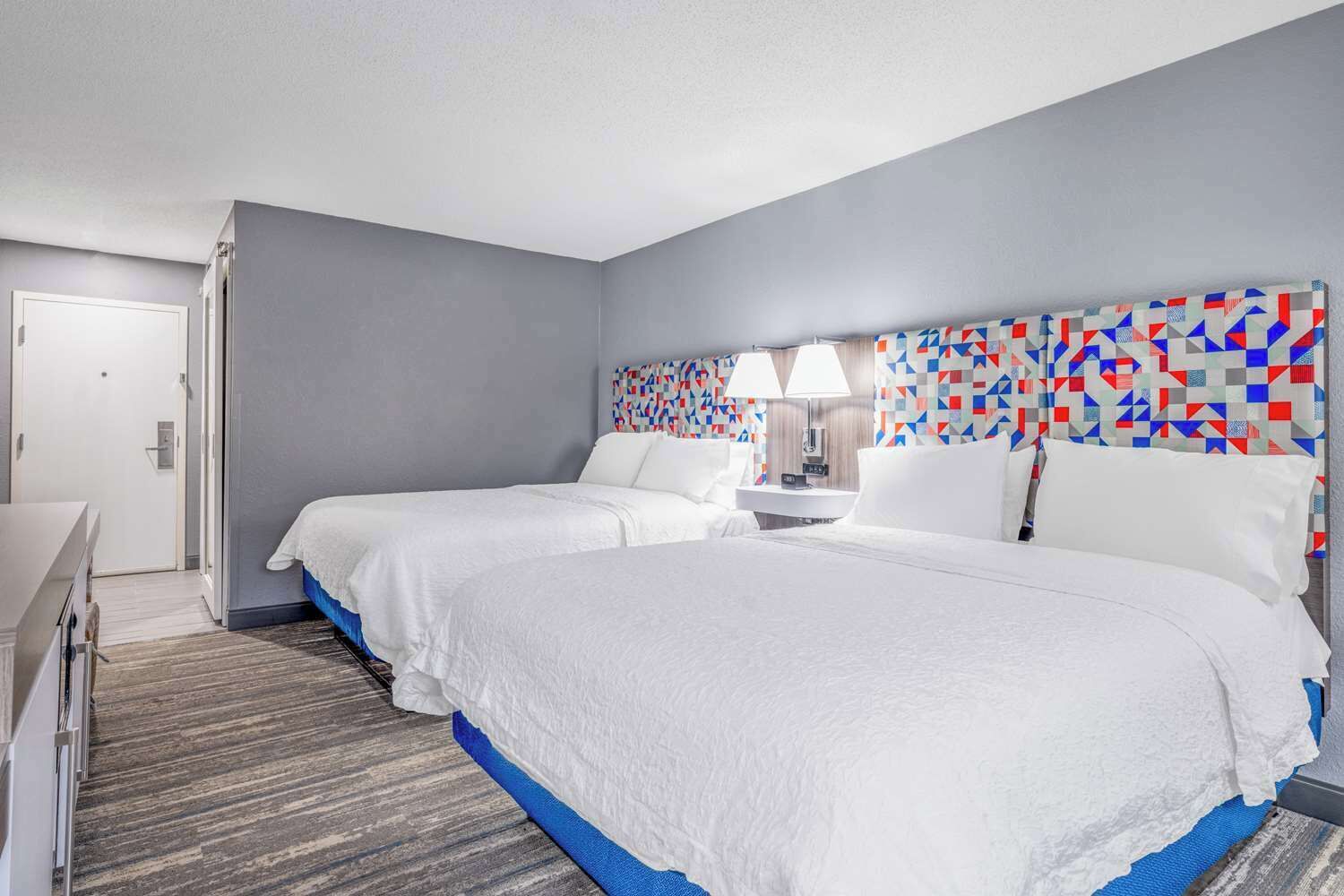 A hotel room with two beds, a desk, a chair, a nightstand, and a patterned headboard.
