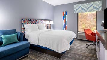 A hotel room with a bed, a blue sofa, a desk with a chair, a window with a view of trees, and a patterned headboard.