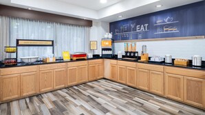 A modern kitchen with wooden cabinets, a black countertop, and a breakfast bar. There are various appliances and breakfast items on the counter. - Hampton Inn Sanford (Sanford)