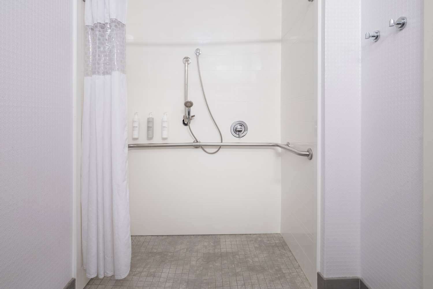 Una ducha con barra de apoyo, pared de azulejos blancos y piso cubierto de baldosas.