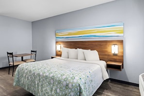 Habitación de hotel con una cama grande, una mesita de noche con lámpara, una silla y una obra de arte colgada en la pared.