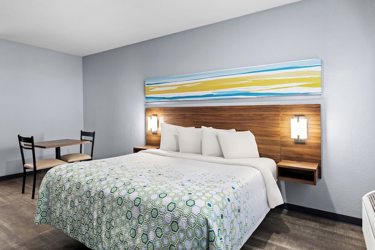 A hotel room with a large bed, a bedside table with a lamp, a chair, and a wall-mounted artwork.