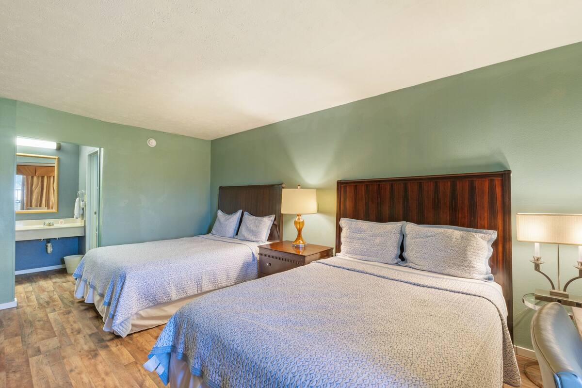 Habitación de hotel con dos camas, cabecera de madera, mesita de noche con lámpara, espejo y baño con lavabo e inodoro.