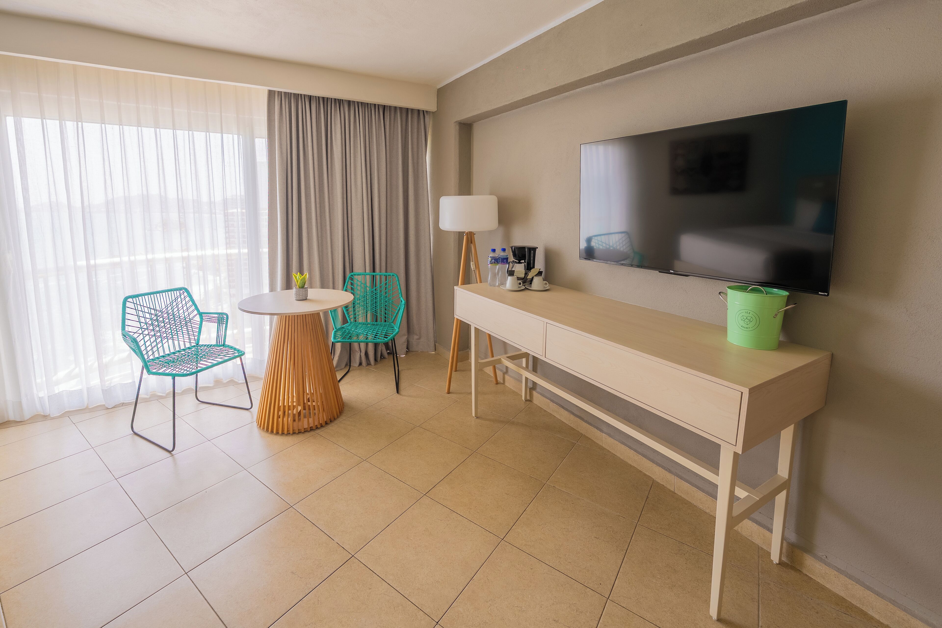 A hotel room with a large flat-screen TV, a wooden desk, two chairs, a small table, and a large window with curtains.