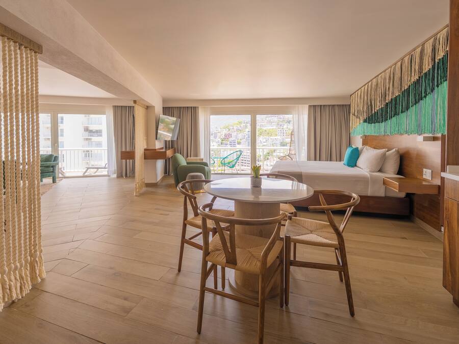 Habitación de hotel con una mesa de comedor redonda, sillas de madera, un sofá y un amplio ventanal con vistas a la ciudad.