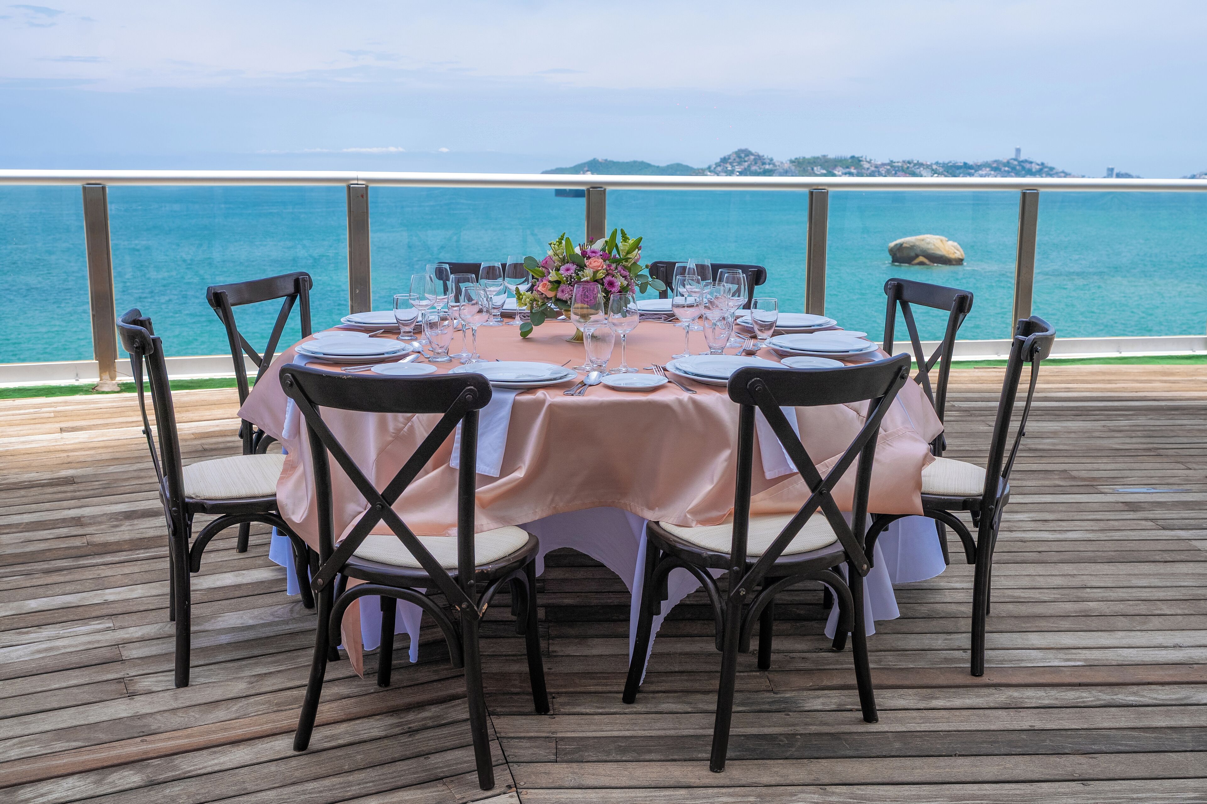 A round table set for six with a pink tablecloth, surrounded by six chairs with cushioned seats, on a wooden deck with a view of the sea.