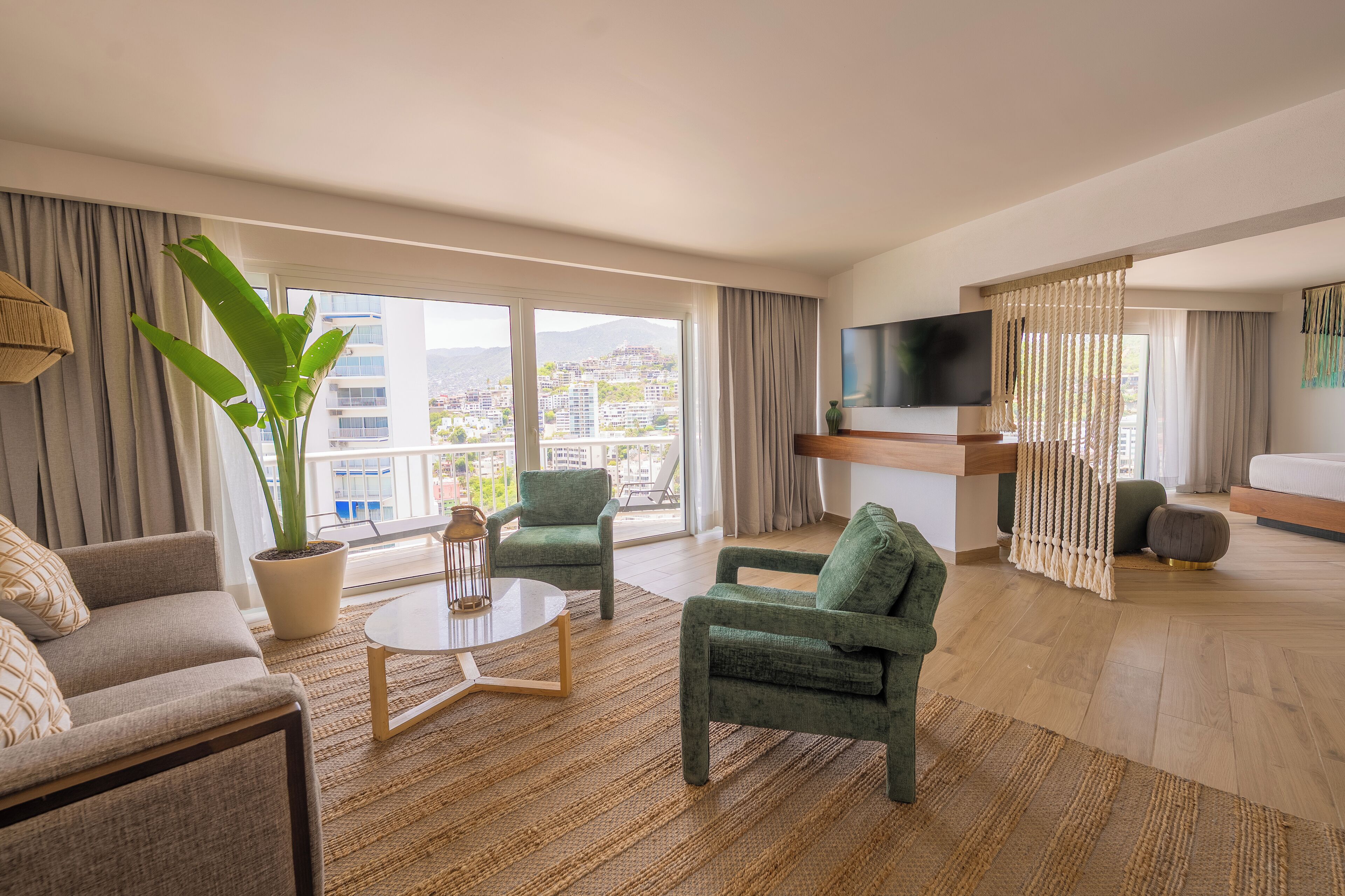 Habitació d'hotel moderna amb un gran finestral, televisió de pantalla plana, un sofà, dues butaques, una taula de cafè i una planta en test.