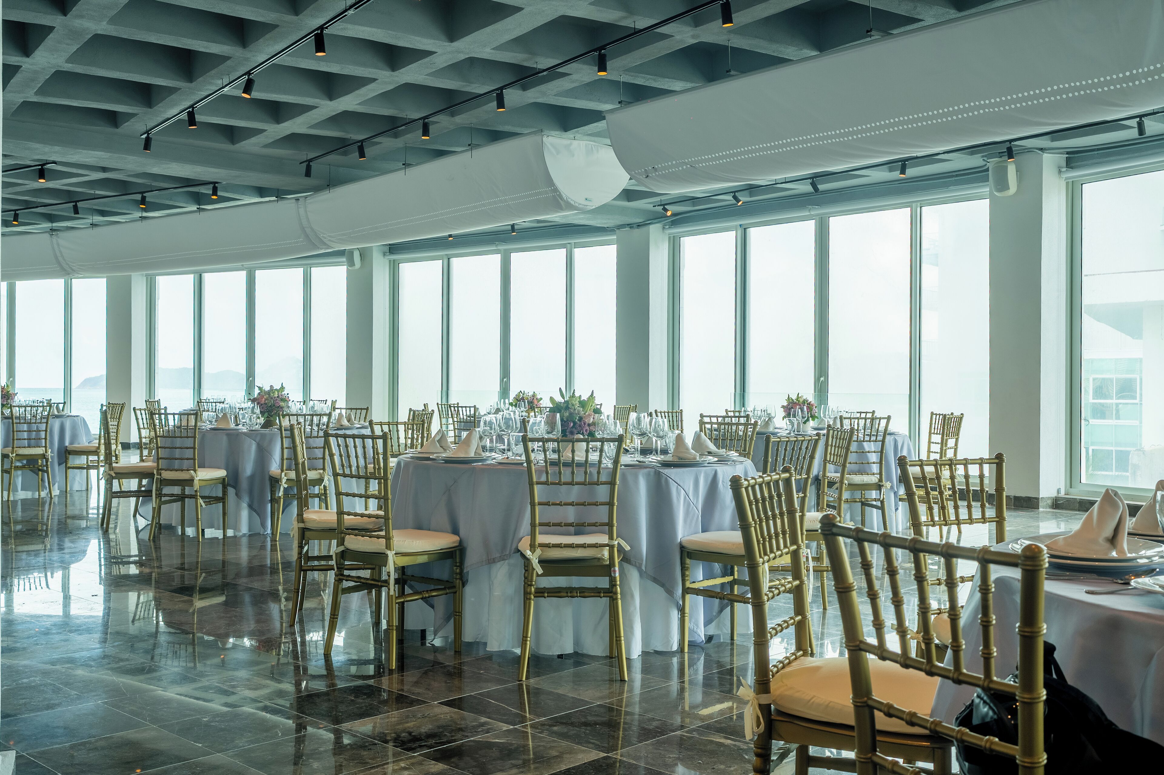 A spacious banquet hall with multiple round tables set for dining, large windows, and a high ceiling with exposed beams.