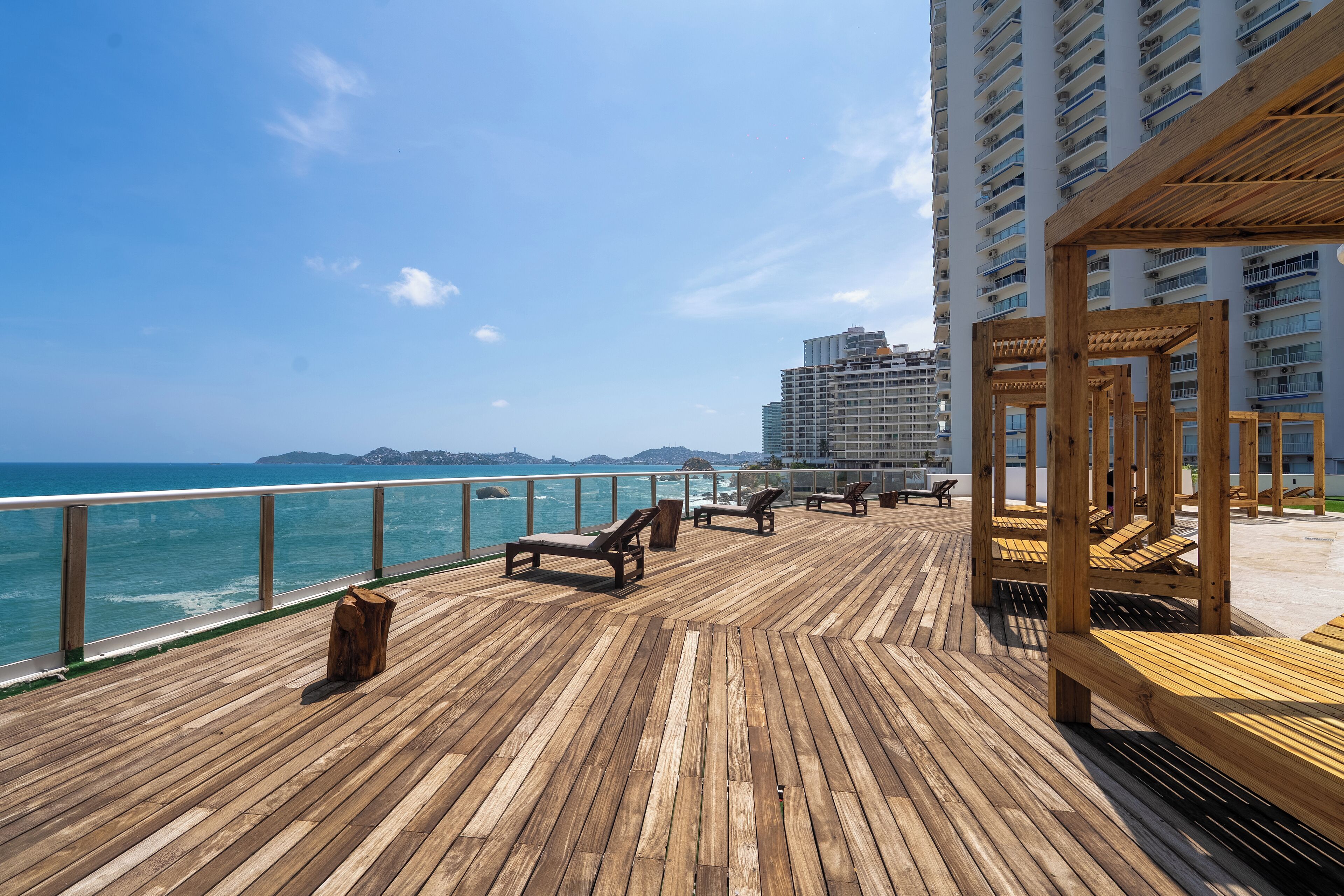 A wooden deck with lounge chairs overlooking the ocean, a wooden pergola, and a high-rise building in the background.
