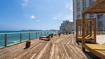 A wooden deck with lounge chairs overlooking the ocean, a wooden pergola, and a high-rise building in the background.