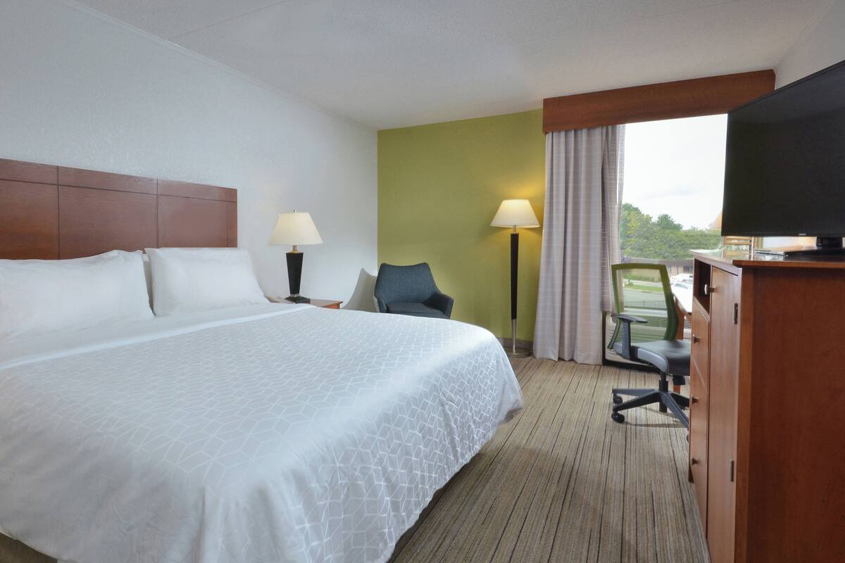 Ein Hotelzimmer mit einem großen Bett, zwei Nachttischlampen, einem Schreibtisch mit Stuhl, einem Fernseher und Blick auf Grünflächen.