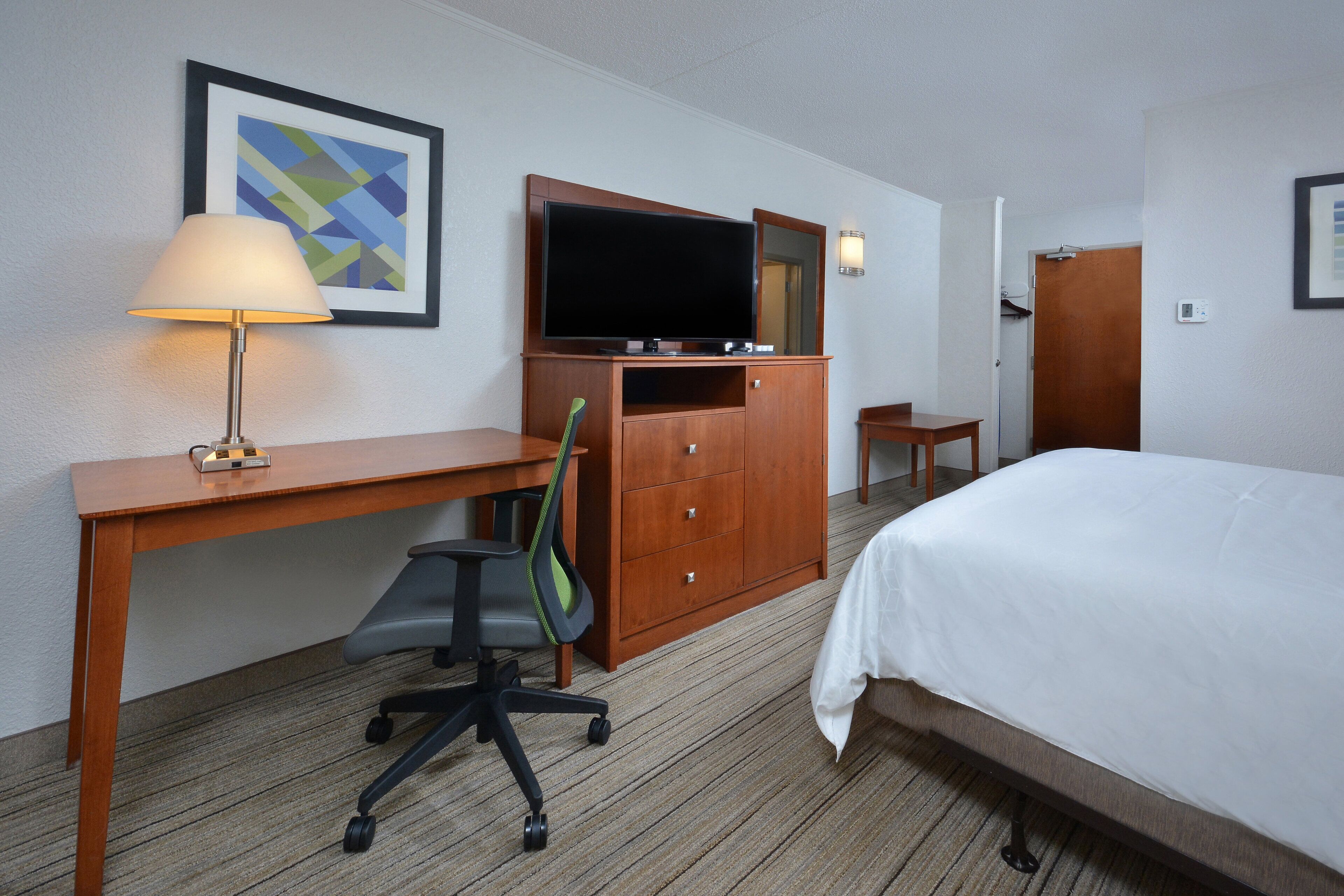A hotel room with a bed, desk, chair, TV, and a framed picture on the wall.