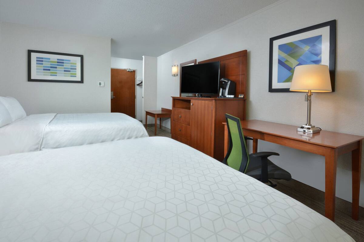 Ein Hotelzimmer mit zwei Betten, einem Schreibtisch, einem Fernseher, einer Lampe und einem gerahmten Bild an der Wand.