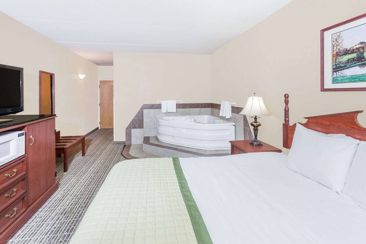 Une chambre d’hôtel comprenant un lit, une télévision, un bureau, une lampe, une tête de lit en bois, un tableau et une salle de bain avec une baignoire et des serviettes.