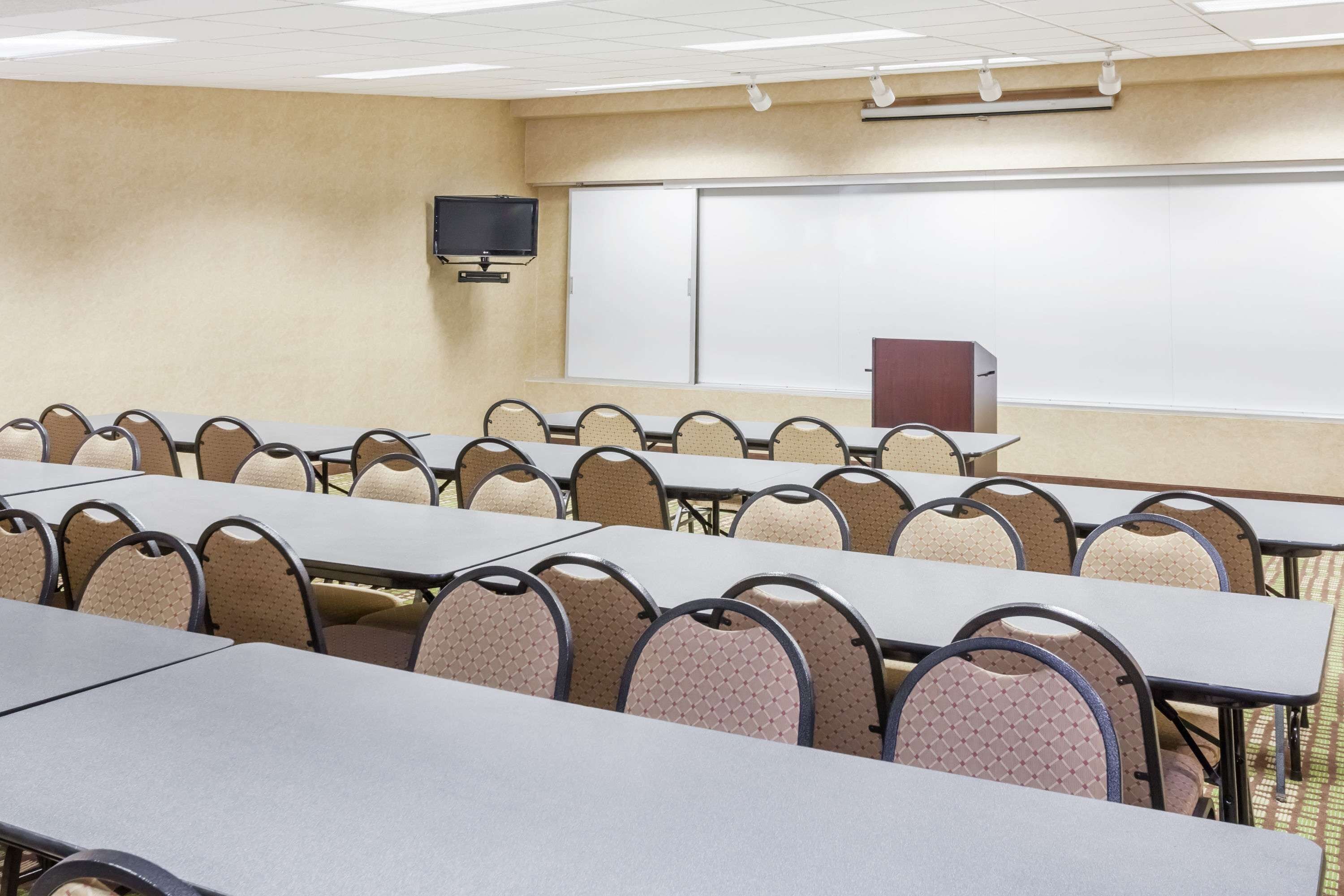 Una sala conferenze con più tavoli e sedie, una lavagna, uno schermo per proiettore e un televisore.