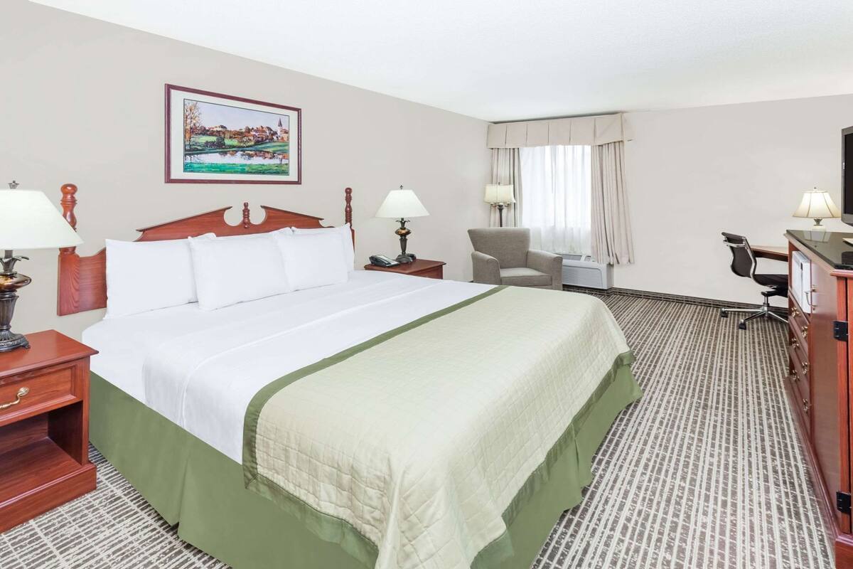 Une chambre d’hôtel avec un grand lit, deux tables de chevet avec des lampes, une chaise, un bureau avec une autre chaise, une fenêtre avec des rideaux et un tableau au mur.