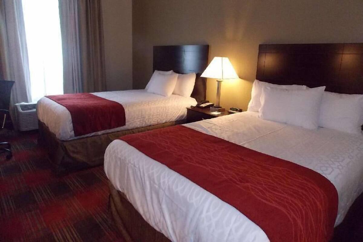 A hotel room with two beds, a nightstand with a lamp, a chair, and a window with curtains.