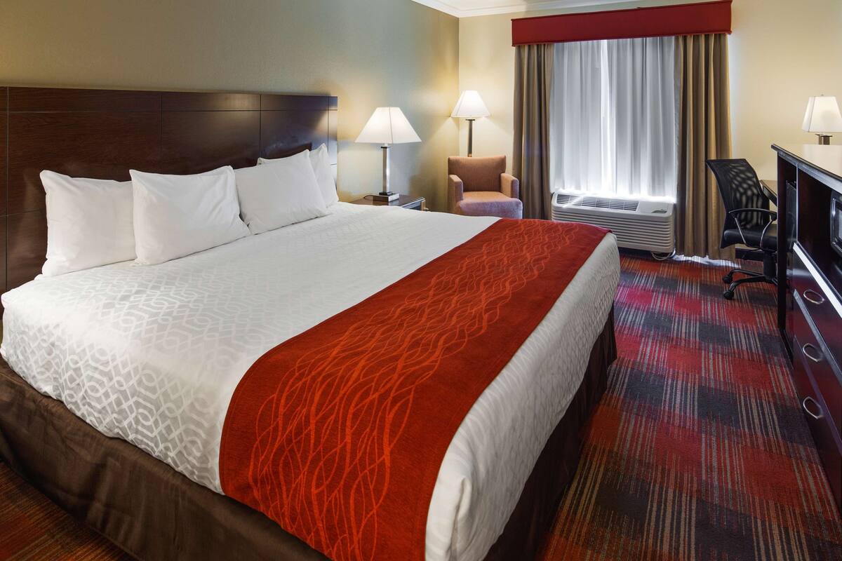 A hotel room with a large bed, two bedside lamps, a chair, a desk with a television, and an air conditioning unit.