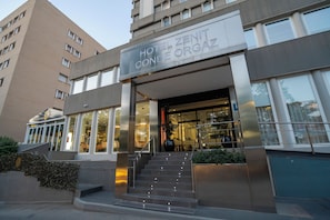 A hotel entrance with a sign reading 'HOTEL ZENIT CONDE ORGAZ,' a staircase leading up to the entrance, and a large window on the right side. - Hotel Zenit Conde de Orgaz (Madrid)
