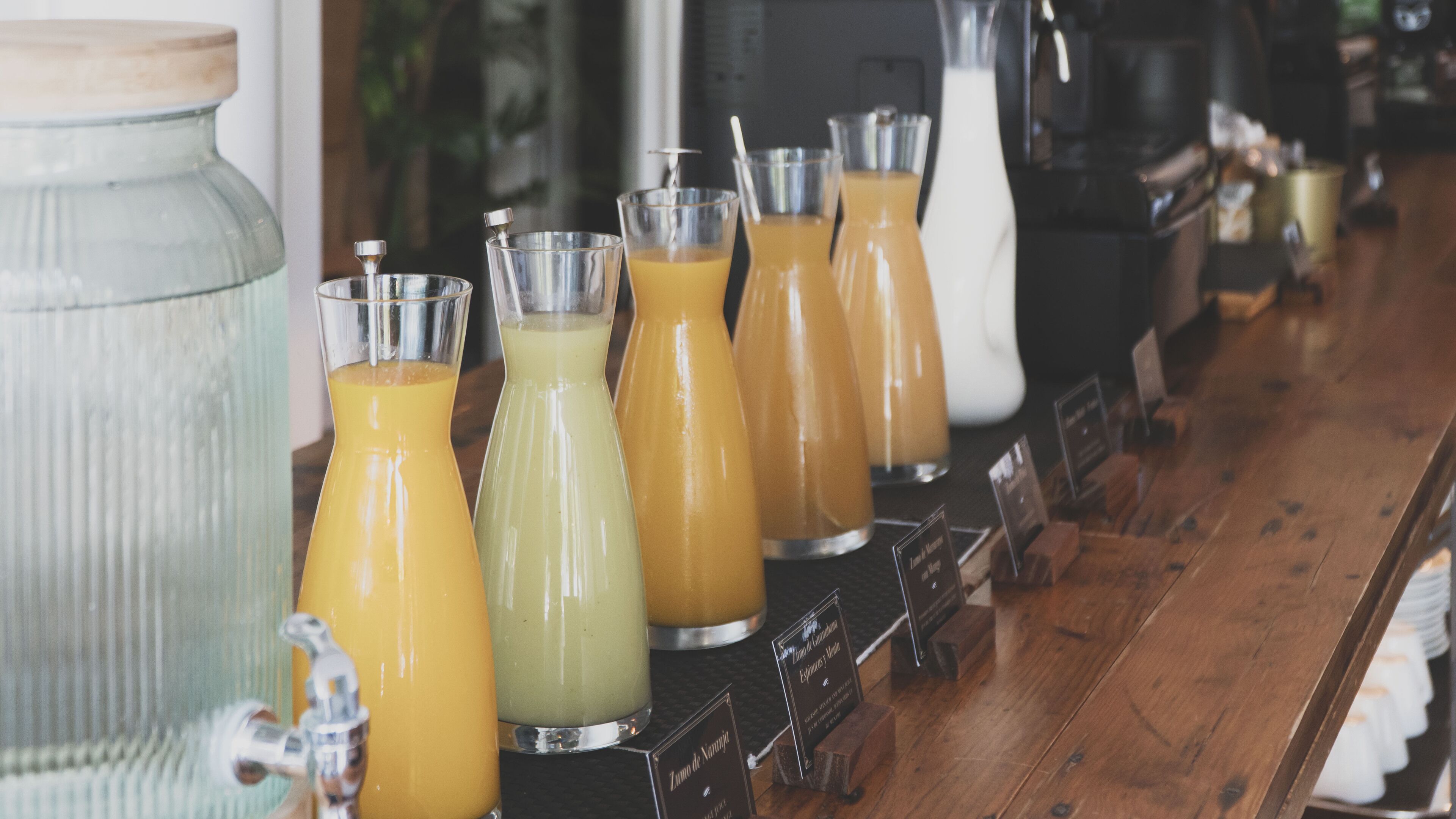 Una zona de bar con diversas bebidas en vasos y botellas, un recipiente de vidrio grande y una barra de madera.
