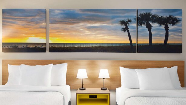A hotel room with two beds, a wooden headboard, a nightstand with two lamps, and three wall-mounted paintings depicting a sunset and palm trees. - Days Inn by Wyndham Kingsland Naval Base I-95 (Kingsland)