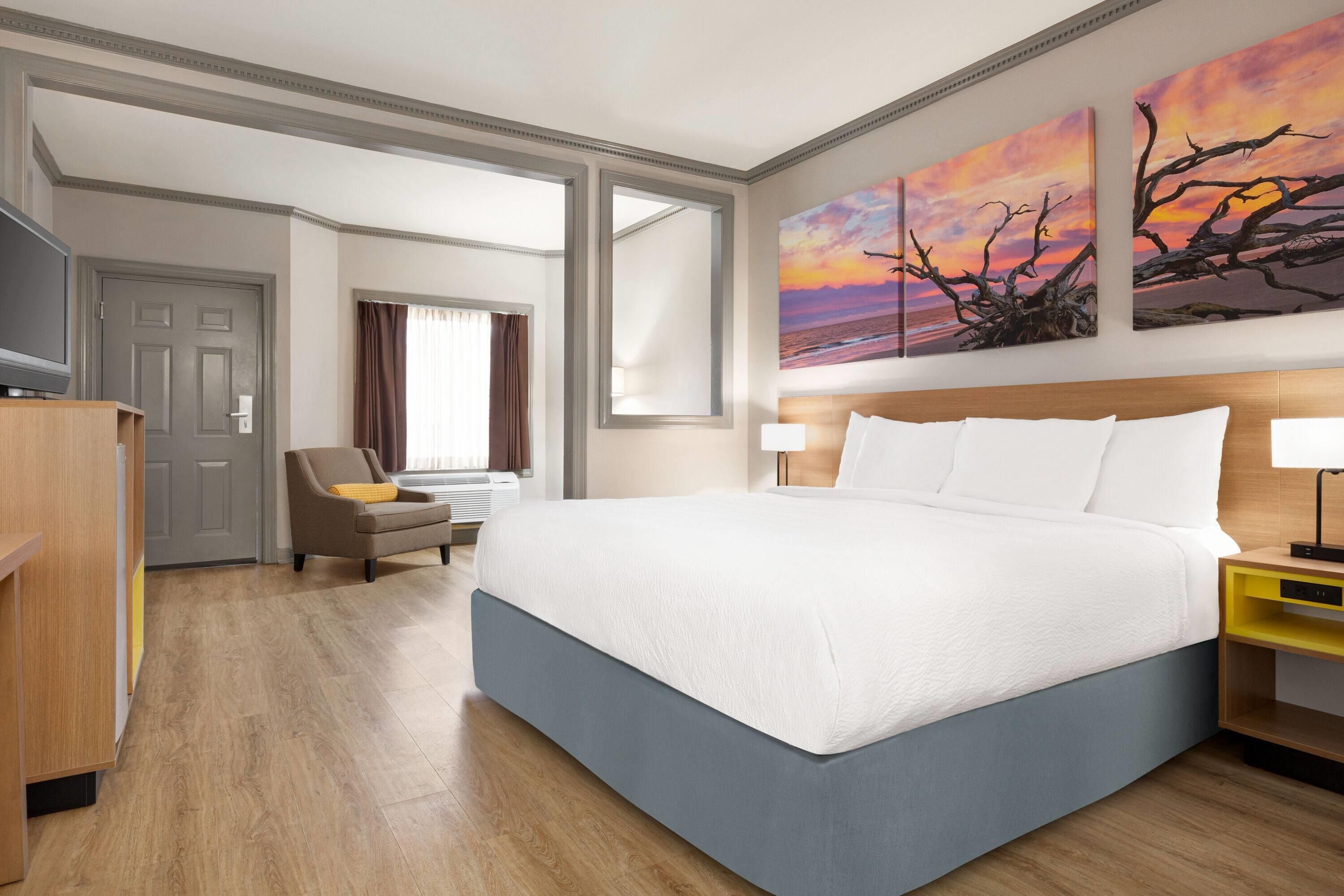 A hotel room with a large bed, a desk, a chair, a television, and a painting of a sunset.