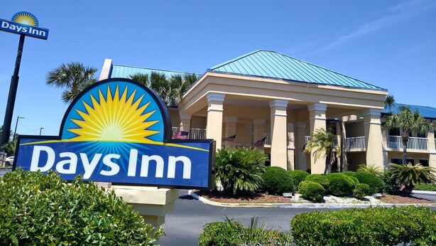 A Days Inn sign with a sun logo, a building with a blue roof, and palm trees. - Days Inn by Wyndham Kingsland Naval Base I-95 (Kingsland)