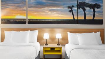 Habitación de hotel con dos camas, una mesita de noche con lámparas y una pared con tres cuadros de atardecer.