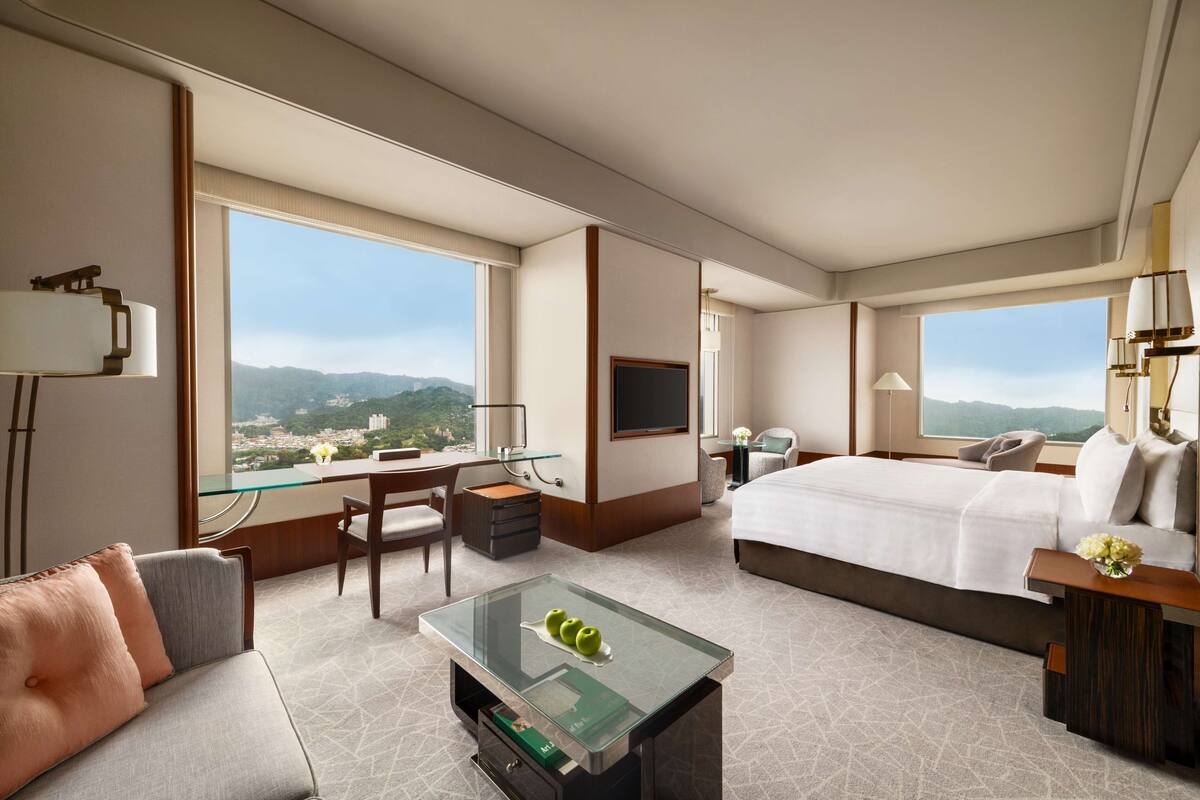 A hotel room with a large bed, a desk with a glass top, a sofa, and a view of the mountains.