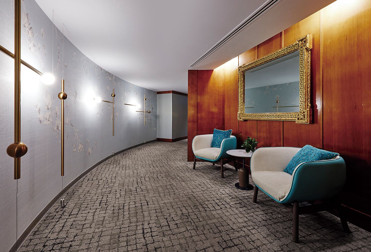 A hallway with a patterned floor, two armchairs with blue cushions, a small round table, and a large mirror with a decorative frame.