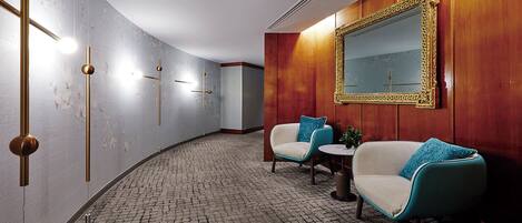A hallway with a patterned floor, two armchairs with blue cushions, a small round table, and a large mirror with a decorative frame.