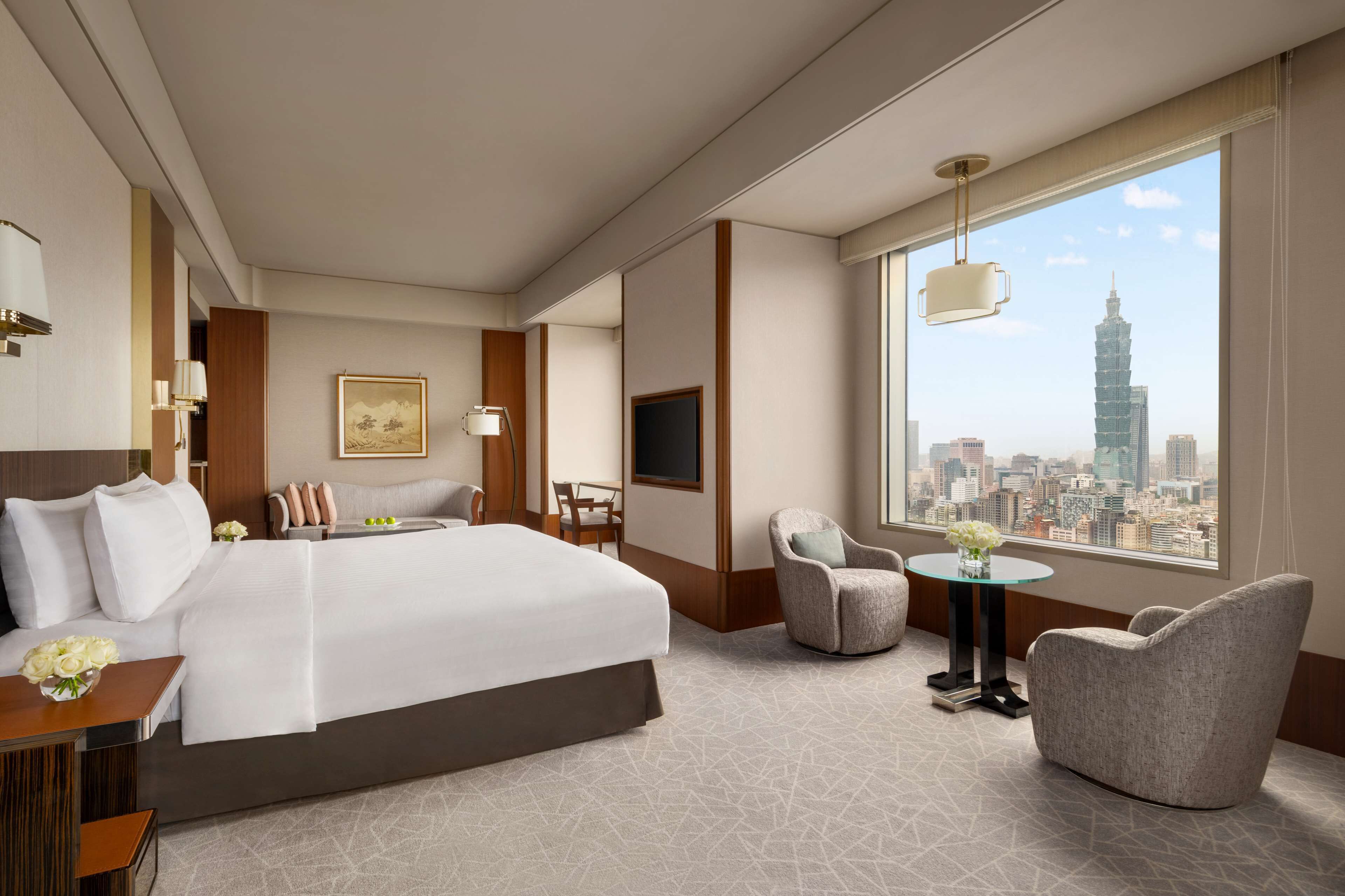 Una habitación de hotel con una cama grande, un escritorio, una silla, una mesita con flores y vistas a un paisaje urbano que incluye un edificio alto.