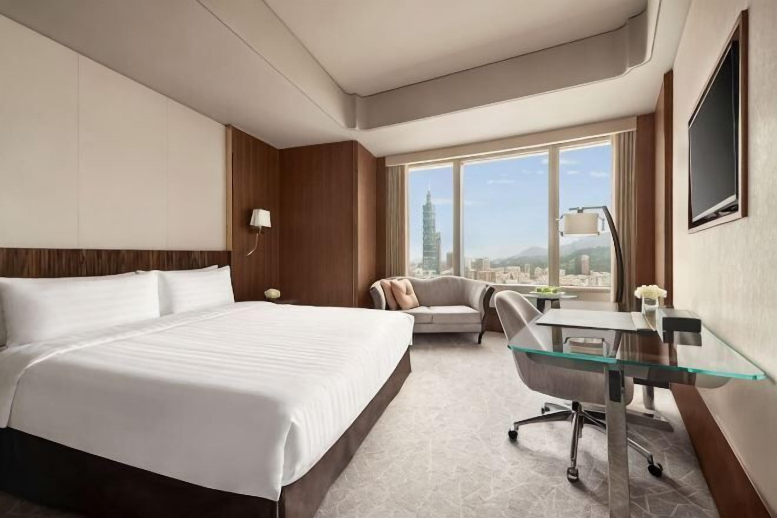 Quarto de hotel com uma cama grande, uma escrivaninha, uma cadeira e vista para a paisagem urbana.