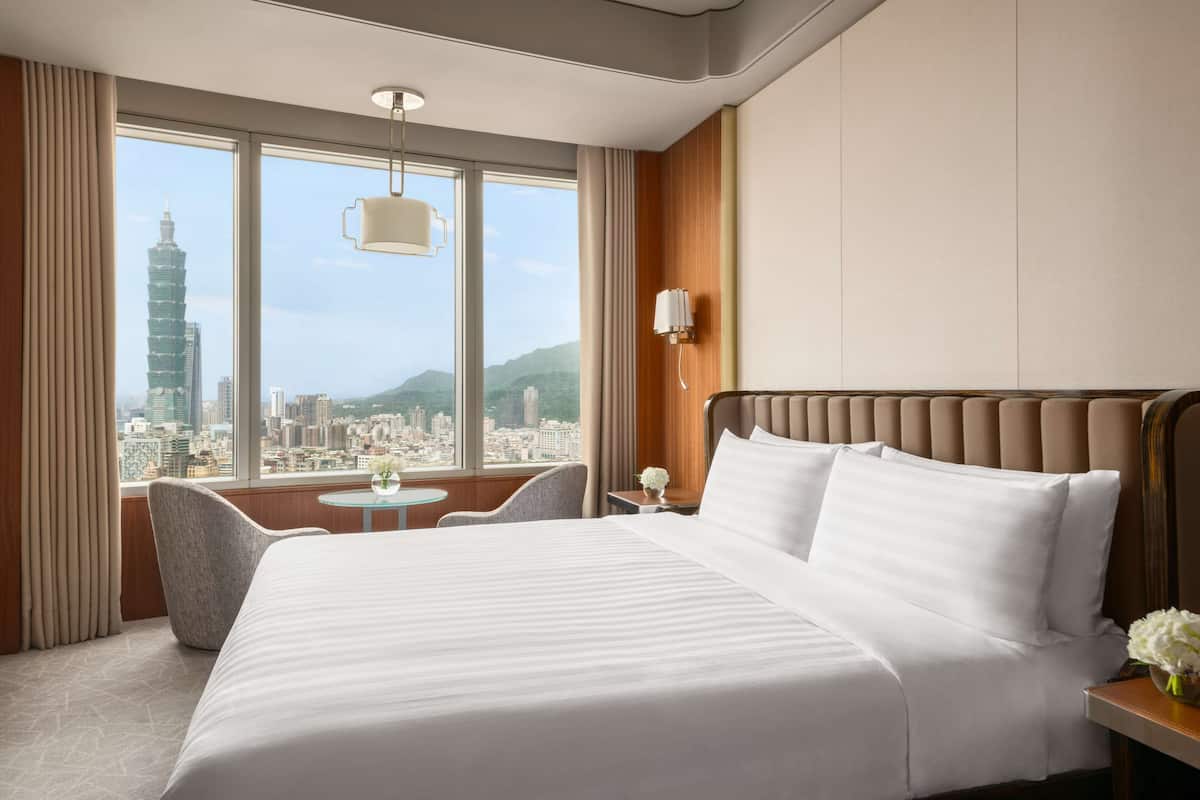 A hotel room with a large bed, a small table, a chair, and a view of a cityscape through large windows.
