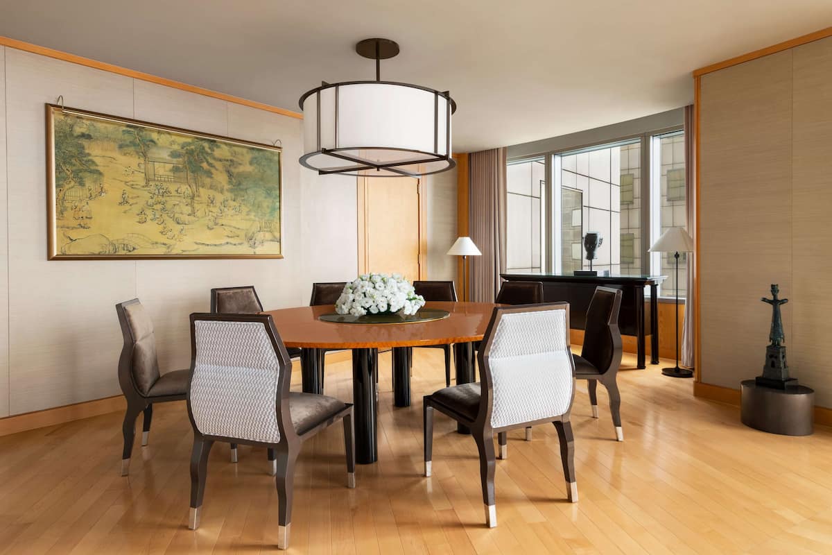 A dining area with a round table, six chairs, a chandelier, a large framed artwork, and a window with curtains.