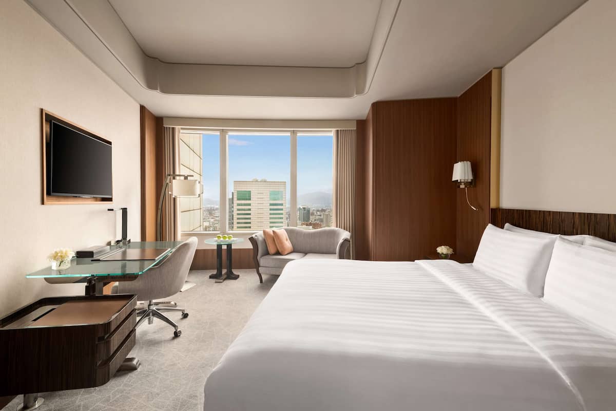 A hotel room with a large bed, a desk with a glass top, a chair, a small table with a vase, and a view of the city through large windows.
